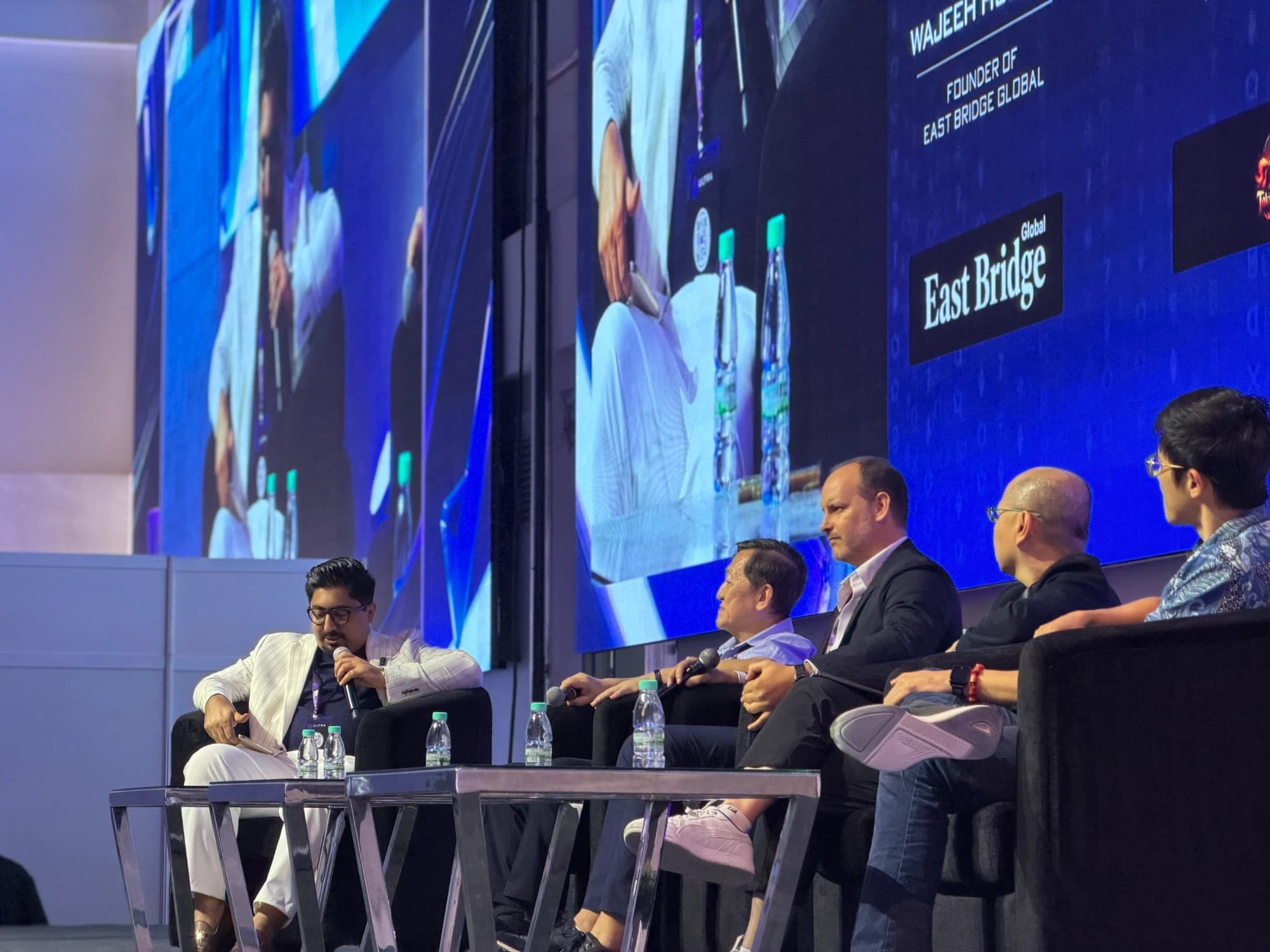 Wajeeh Hussain, microphone in hand, moderating a panel of four on stage at ULTRA Web3 Festival 2026 in Kuala Lumpur, with his name and the East Bridge Global logo on the screen behind.
